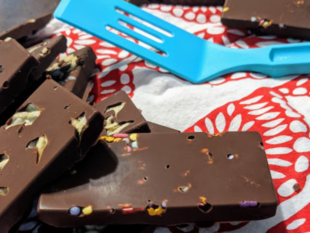 A stack of homemade chocolate bars filled with sprinkles and potato chips with a blue spatula in the background. 
