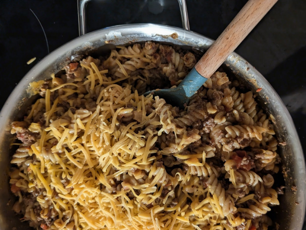 A stainless steel skillet filled with cheesy ground beef, rotini pasta, and corn, topped with melted cheddar cheese.